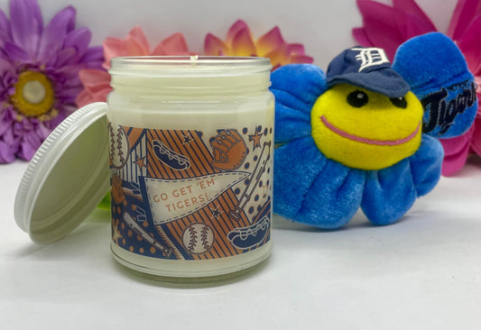 Hand-poured candle in clear glass with Detroit Tigers label & white lid resting beside it, next to a flower with smiley face.