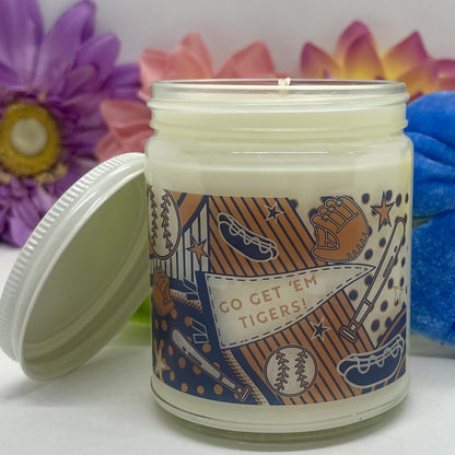 Hand-poured candle in clear glass with Detroit Tigers label with white lid resting beside candle and floral background.