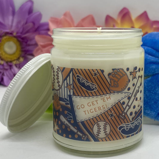 Hand-poured candle in clear glass with Detroit Tigers label with white lid resting beside candle and floral background.