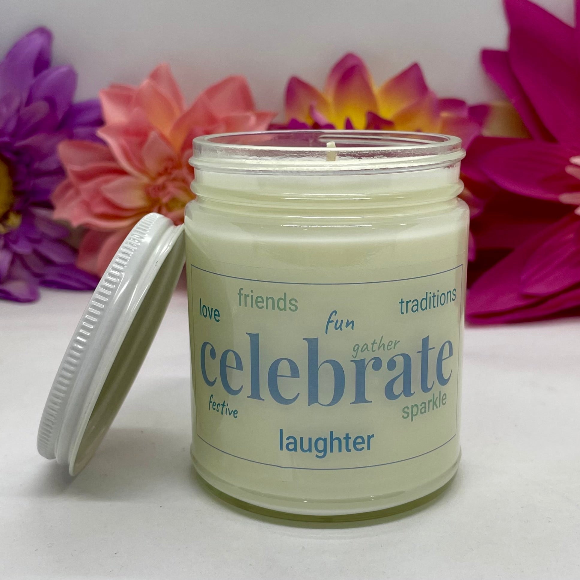 A hand poured candle with label that says Celebrate & other positive words, white lid resting next to it & floral background.
