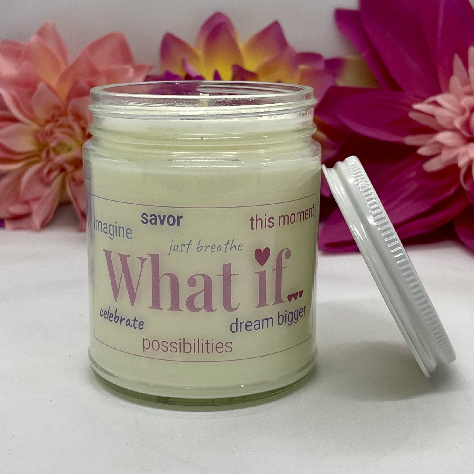 A hand poured candle with label that says What If & other positive words, white lid resting next to it & floral background.