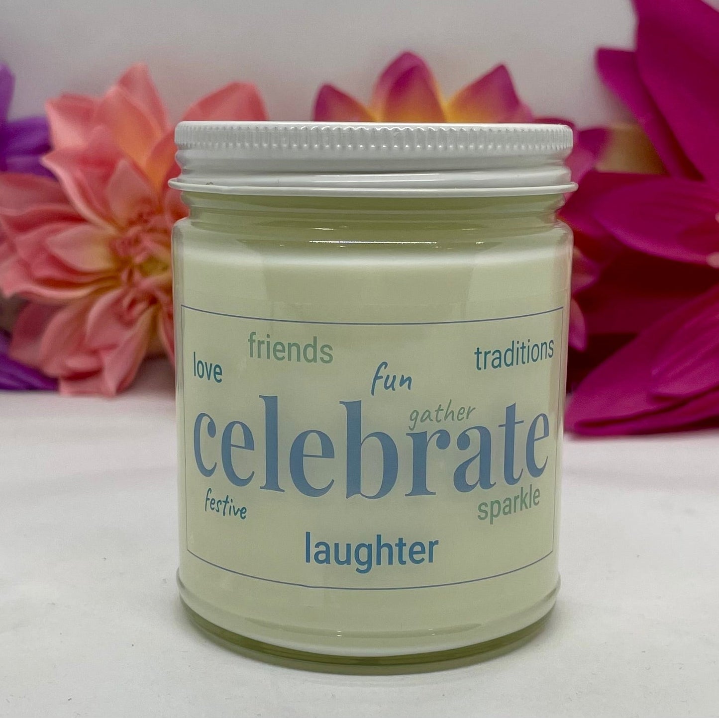 A hand poured candle with label that says Celebrate and other positive words, with white lid and bright floral background.