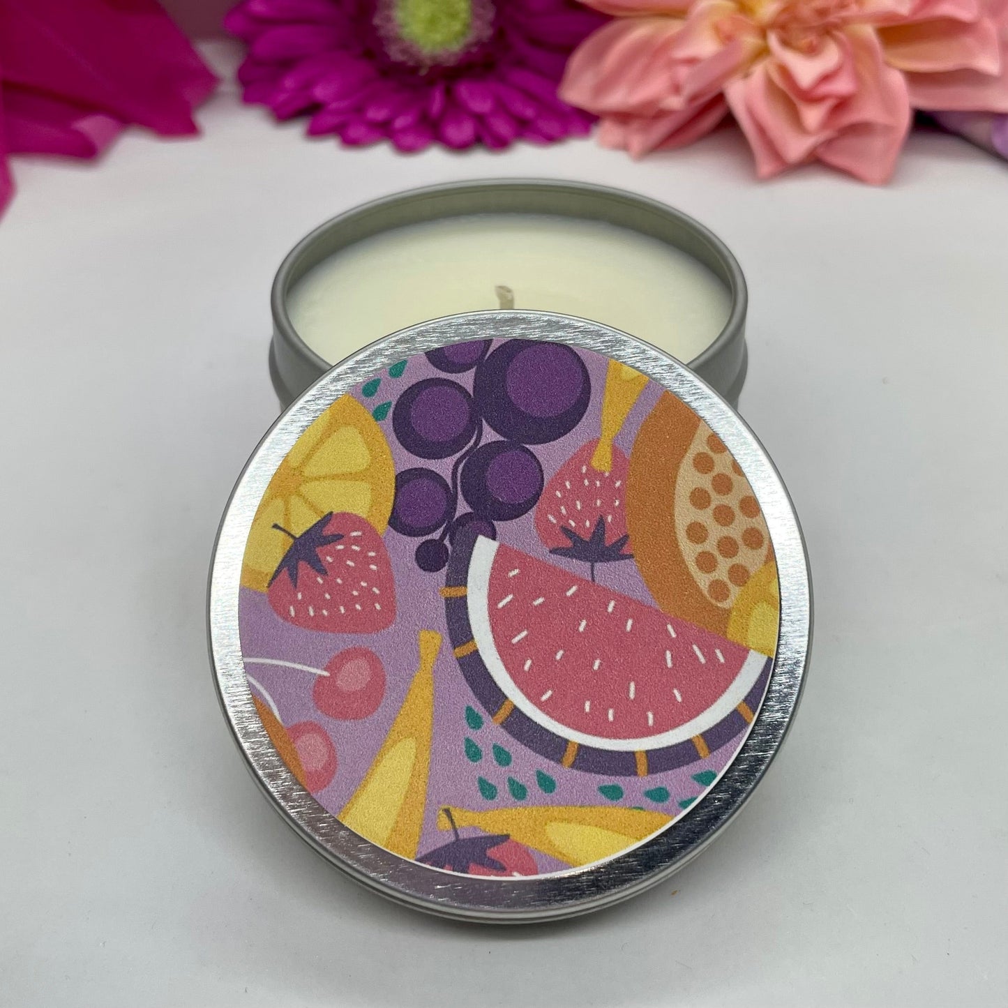 Hand poured candle in silver tin with colorful fruit label on lid with lid resting in front, with bright floral background.