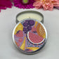 Hand poured candle in silver tin with colorful fruit label on lid with lid resting in front, with bright floral background.