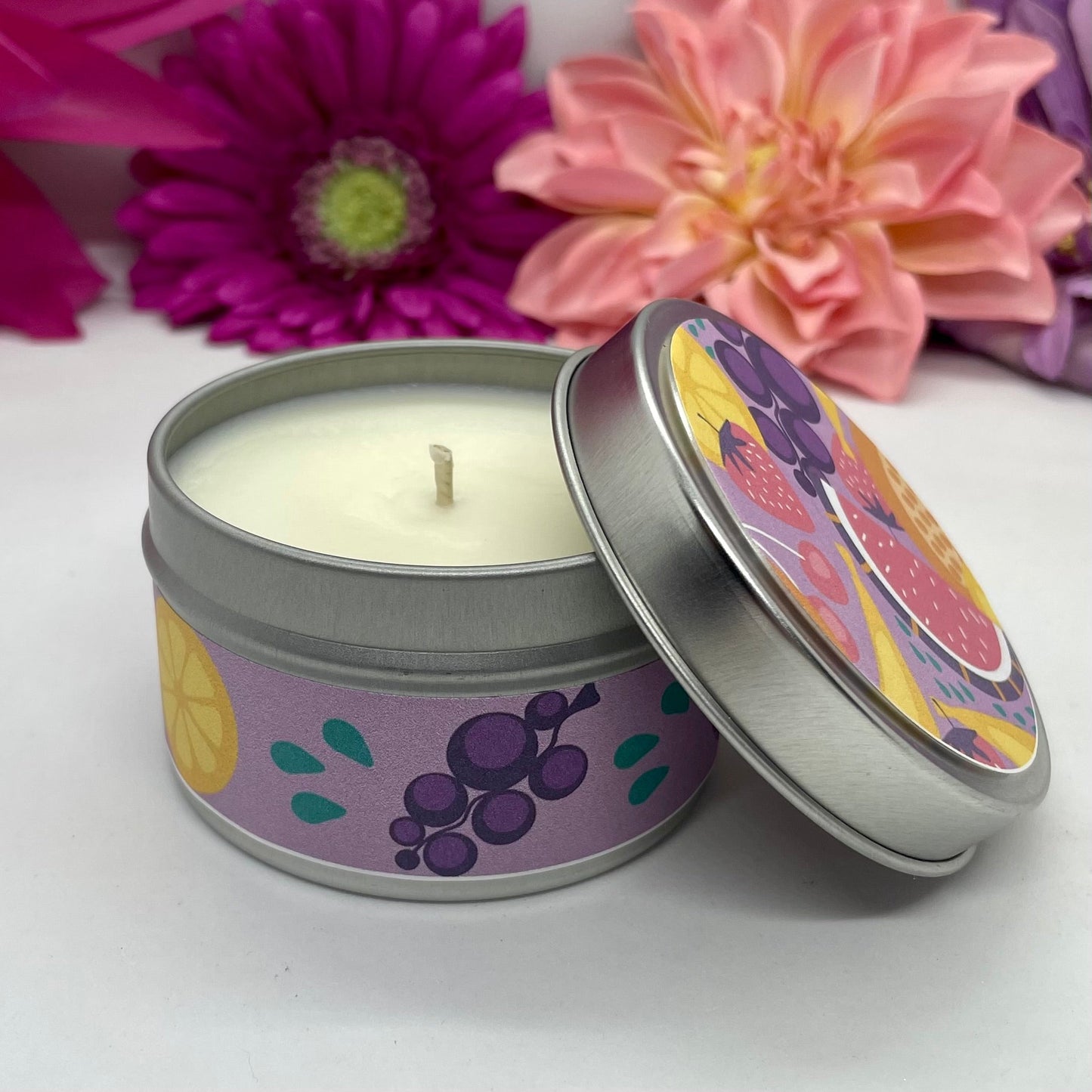 Hand poured candle in silver tin with colorful fruit labels with lid resting to the side, with bright floral background.