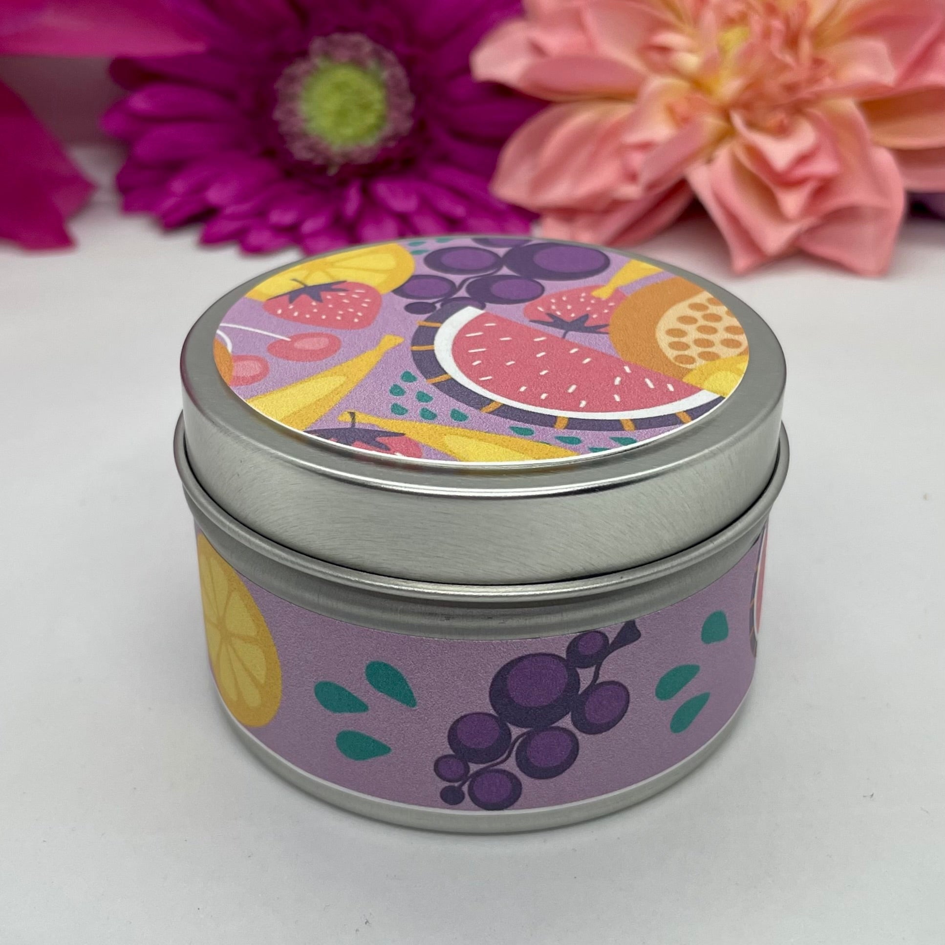 Hand poured candle in silver tin with colorful fruit label on lid and body of the tin, with bright floral background.